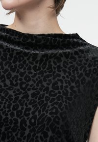 Osoba v černém sametovém topu se subtilním vzorem leopardího tisku a širokým lodičkovým výstřihem, zkráceným pod bradou.
