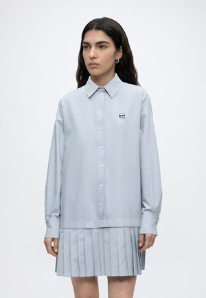 Femme aux longs cheveux foncés portant une chemise bleu clair boutonnée avec un petit logo brodé et une jupe plissée assortie.