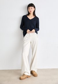 Tume sinise pika varrukaga V-kaelusega kootud jakk, millel on mugav lõige, koos kreemjate laia säärega pükste ja pruuni seemisnahast platvormkingadega neutraalsel taustal.