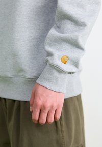 Sweatshirt gris avec des poignets côtelés, arborant un petit logo jaune brodé sur la manche. Matière douce, coupe décontractée et design minimaliste.