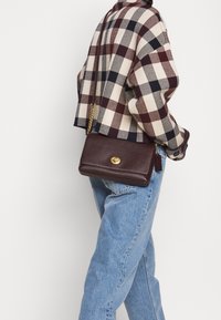 Ruskea nahkainen käsilaukku, jossa on kultainen lukko ja ketjuhihna. Laukku on suorakulmion muotoinen ja sileäpintainen, käytetty ruudullisen takin alla.