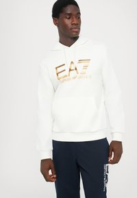 Sweatshirt branca com logótipo impresso a dourado a dizer "EA7 Emporio Armani", apresentando um bolso frontal e capuz com cordão. Combinada com calças de moletom azul-marinho.