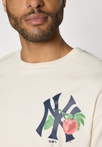 Cream-colored cotton t-shirt featuring a large navy blue NY logo with a red apple and green leaves design. Short sleeves, round neckline.