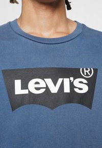 Gros plan d'un t-shirt bleu à col rond avec un grand logo Levi's noir et blanc sur la poitrine, porté par une personne aux dreadlocks courtes.
