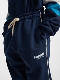 Person iført marineblå hummel sportbukser med snøre i taljen og hænder i lommerne, der viser en del af en matchende jakkeærme.