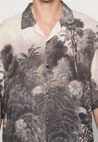 Uomo che indossa una camicia beige con bottoni, decorata con una stampa tropicale nera che raffigura alberi e fogliame.