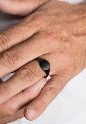 Juodas metalinis žiedas su lygiu, suapvalintu paviršiumi, dėvimas ant rankos su trumpais, šviesiais plaukais ir matoma odos tekstūra. Paprastas, modernus dizainas.