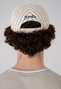 Beige caps med marineblå striper, med "Berlin" brodert i skrivestil på baksiden. Bæres med en lys grå, ribbestrikket genser.