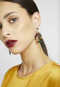 Pendientes llamativos y coloridos con un diseño ovalado de acrílico, que cuentan con un anillo dorado texturizado en la parte superior y una mezcla de patrones vibrantes.