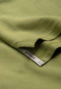 Gros plan sur un tissu vert texturé avec des coutures visibles et une étiquette blanche de vêtement montrant partiellement le nom de la marque.