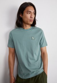 Tricou de culoare teal cu maneci scurte, fabricat din bumbac moale, având un mic logo alb Puma pe un petic pătrat. Guler simplu de tip crew și croială lejeră.