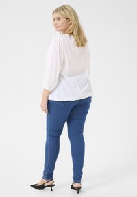 Bl blouse blanc texturé avec des manches bouffantes, associé à un jean skinny bleu et des mules à talons noirs, présentant une tenue décontractée.