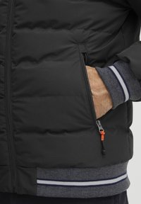 Svart pufferjakke med en glidelåslomme med oransje trekk, og grå ribbestrikkede mansjetter med blå og hvite striper. Teksturert stoff, isolert design.