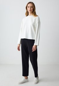 Donna in piedi che indossa una maglietta bianca a maniche lunghe con piccoli punti, pantaloni neri dritti e sneakers bianche, contro uno sfondo chiaro e semplice.