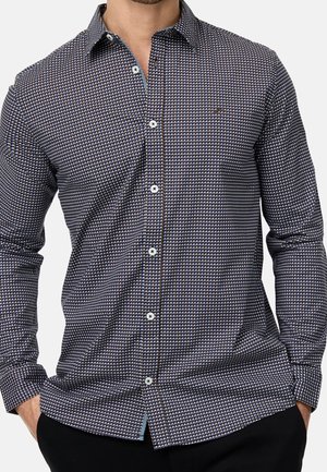 Mężczyzna ubrany w ciemnoniebieską, wzorzystą koszulę na guziki z długim rękawem, ozdobioną małymi białymi i jasnoniebieskimi geometrycznymi kształtami.