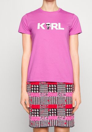 Vrouw draagt een felroze T-shirt met de tekst "KARL" en een klein cartoonfiguurtje met een zonnebril, gecombineerd met een rode en roze rok met patroon.