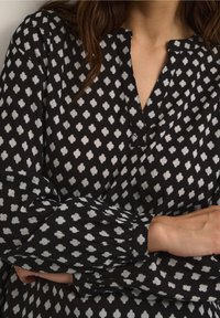Frau in einer schwarzen Bluse mit langen Ärmeln und weißem geometrischem Muster, kragenlosem Ausschnitt und knöpfbaren Manschetten, die die Arme verschränkt hat.