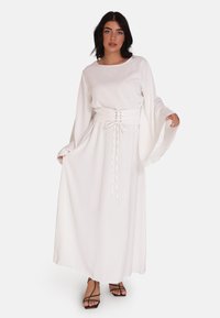 Robe longue blanche avec une taille cintrée, des manches larges et une texture lisse. Présente un détail à nouer à la taille et est associée à des sandales noires à lanières.