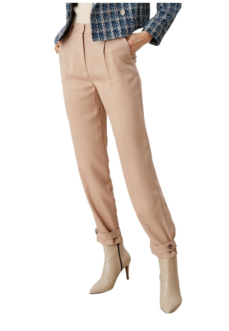 Beige skräddarsydda byxor med veck och uppvikta fållar, kombinerat med nakna högklackade stövlar. Tyget ser slätt ut med en strukturerad design.
