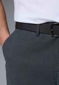 Pantalon gris texturé avec un motif subtil, arborant une ceinture en cuir noir et une chemise blanche partiellement visible en arrière-plan.