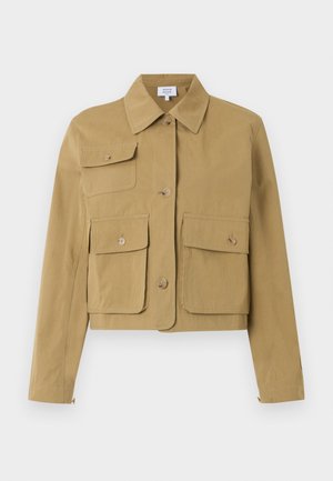 Vrouw met zwarte zonnebril, een beige cropped jas met grote zakken, een wit topje, lichtblauwe high-waist wijde jeans en zwarte platte schoenen.