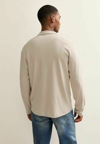 Camisa de polo de manga larga, de color beige claro, con una textura suave y un dobladillo redondeado. Combinada con unos pantalones vaqueros azules. Vista desde atrás.