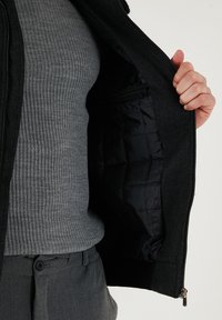 Črn volnen plašč s črno podlogo iz prešite tkanine, z zapiranjem na zadrgo in dvema notranjima žepoma. Zunanjost ima mehko teksturo in kontrastni ovratnik.