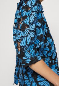 Blue floral lace top featuring intricate black embroidery, textured design, short sleeves, and a flowy silhouette.