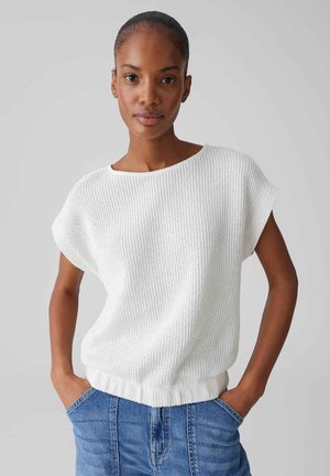 Frau mit kurzen Haaren, die ein weißes, gestricktes ärmelloses Oberteil und blaue Jeans trägt, steht mit den Händen in den Taschen vor einem einfarbigen Hintergrund.