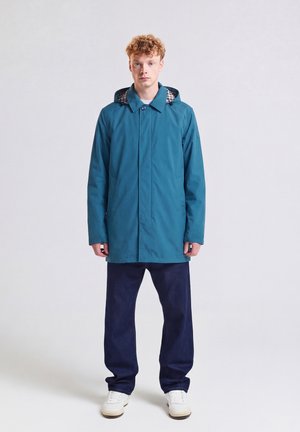 Young man with curly hair wearing a teal hooded coat, dark blue jeans, and white sneakers standing against a plain light background.