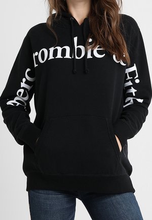 Femme portant un sweat à capuche noir avec le logo blanc « Abercrombie & Fitch » sur la poitrine et les manches, les mains dans la poche avant, associé à un jean bleu.