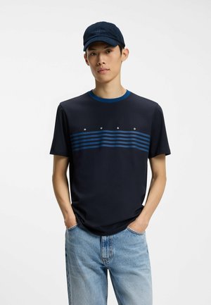 Homme portant une casquette bleu marine, un T-shirt bleu marine à rayures bleues avec le texte "HUGO" et un jean bleu clair, debout les mains dans les poches.