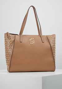 Torebka tote w kolorze brązowym z metalowymi dodatkami, złotym logo i dwiema uchwytami. Gładka tekstura z kontrastującymi, ćwiekowanymi detalami po bokach.