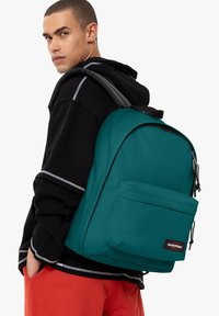 Zaino in nylon color teal con tasca frontale, spallacci imbottiti e patch con logo. Il modello indossa una felpa nera e pantaloni arancioni.