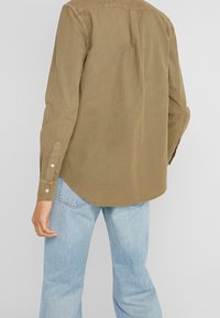 Khaki košile s dlouhým rukávem a zakřiveným lemem, vyrobená z hladkého materiálu. Obsahuje zadní skládku pro pohodlnější pohyb.