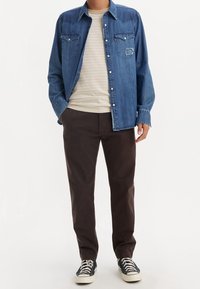 Chemise en denim avec deux poches poitrine, bleu clair, portée sur un t-shirt rayé crème, associée à un pantalon marron et des baskets noires.