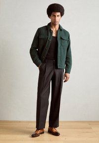 Giacca verde texturizzata con due tasche sul petto, camicia nera aderente, pantaloni neri su misura e scarpe in pelle marrone. Pavimento in legno naturale.