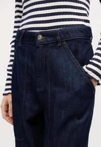 Mørkeblå denim bukser med kontrastfarget oransje søm, fem-lommers design og en metallknapp lukking i livet. Stripet langermet topp synlig.