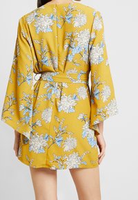 Florales Wickelkleid in Senfgelb mit weißen und blauen Blumen, mit weiten Ärmeln und einem gebundenen Gürtel in der Taille. Glatte Textur.