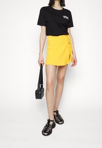 T-shirt noir avec broderie florale, mini jupe côtelée jaune avec boutons latéraux, sac bandoulière noir et sandales noires à lanières.