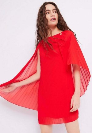 Donna con lunghi capelli ricci che indossa un vestito rosso con maniche a mantella trasparenti, tenendo da una parte il mantello fluente.