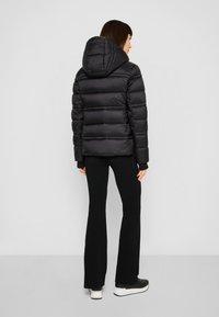 Zwarte pufferjas met horizontaal doorgestikt ontwerp, capuchon, elastische manchetten en voorzakken. Draagt zwarte aansluitende broek en sneakers.