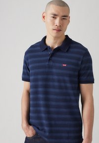 Polo de culoare albastru marin cu dungi, confecționat din bumbac, având un guler clasic, placket cu trei nasturi și un mic logo Levi's roșu pe piept.