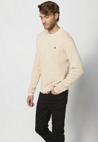 Beige geribbelde trui met lange mouwen en een ronde halslijn, met een klein marineblauw logo op de borst, gecombineerd met zwarte slim-fit broek.