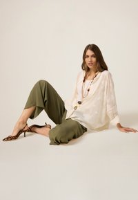 Blusa bianca leggera e trasparente con motivo floreale, abbinata a pantaloni larghi verde oliva e scarpe con punta a forma di leopardo. Collana lunga inclusa.