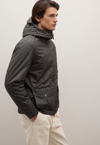 Giacca con cappuccio grigio scuro con cerniera, dotata di due tasche frontali e cappuccio pieghevole, realizzata in tessuto a texture liscia.