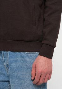 Δεξί χέρι ενός ατόμου με σκούρο καφέ φούτερ και ανοιχτό γαλάζιο τζιν κατά των plain φόντο.