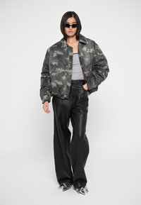 Grijze camouflage bomberjack, aansluitend grijs tanktop, zwarte leren broek en metallic puntige hakken. Zonnebril maakt de look compleet.