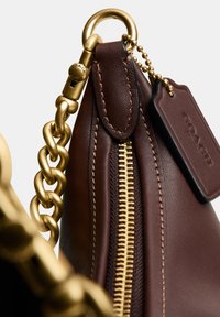 Dark brown leather bag corner with gold zipper, chain strap, and a leather tag embossed with "COACH" on a ball chain.