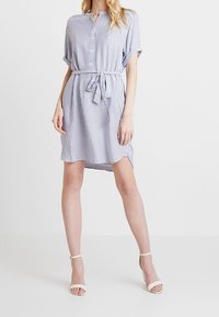 Sukienka w paski w kolorze niebieskim i białym, krótki rękaw, z przodu na guziki oraz dopasowany pasek w talii. Tkanina wydaje się lekka i gładka.